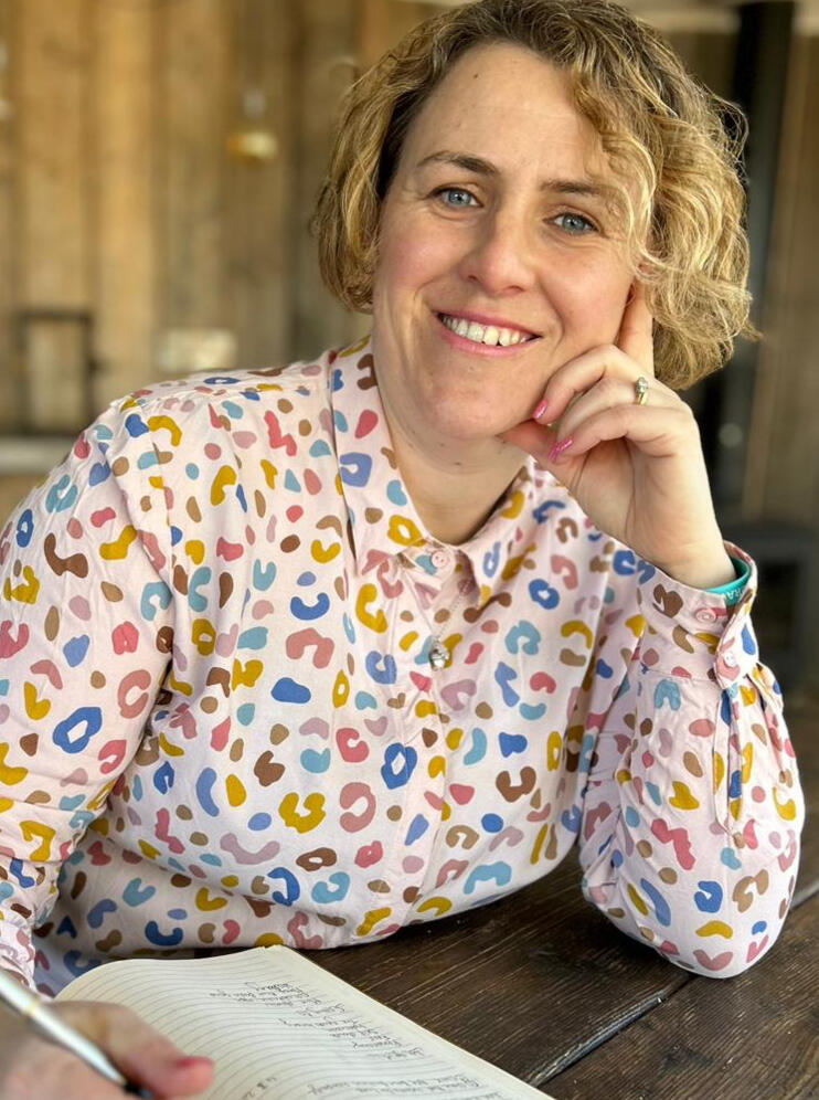 A smiling white woman in her thirties sitting at a wooden table and wearing a colourful leopard print shirt with a pen in her hand.